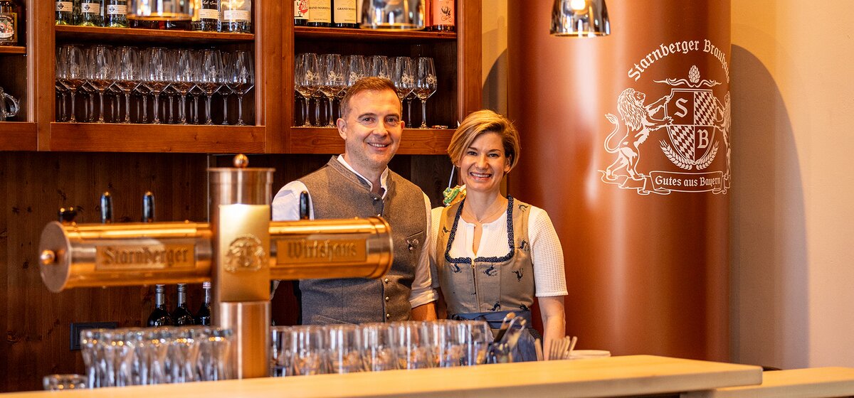 Aleks und Steffi in traditioneller bayerischer Tracht stehen hinter einer Bar mit Biergläsern und Zapfhähnen, im Hintergrund ein großer Tankbier-Kessel mit dem Schriftzug 'Starnberger Brau'.